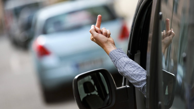 Ein Autofahrer zeigt seinen Mittelfinger einem anderen Verkehrsteilnehmer aus dem Fenster seines Autos. (gestellte Szene)