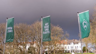 Drei Werder-Fahnen wehen vor dem Weser-Stadion mit Blick auf den Osterdeich mit schwarzen Gewitterwolken am Himmel.