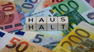 Auf Euronoten liegende Würfel zeigen das Wort "Haushalt" (Symbolbild)
