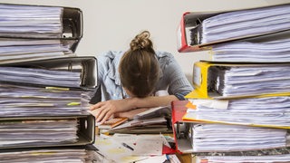 Junge Frau arbeitet am Schreibtisch zwischen Aktenbergen.