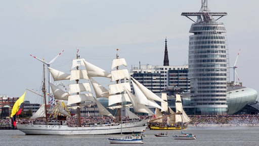 Mehrere Schiffe fahren durch den Hafen in Bremerhaven. Im Hintergrund sind Gebäude und viele Menschen zu sehen.