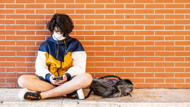 Ein Jugendlicher sitzt an einer Wand, trägt einen Mund-Nase-Schutz und blickt auf sein Smartphone.