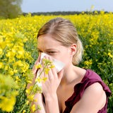 Eine Junge Frau steht in einem gelb blühenden Feld und niest in ein Taschentuch.