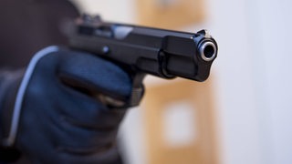 Eine Hand mit Handschuh hält eine Schusswaffe in die Kamera.