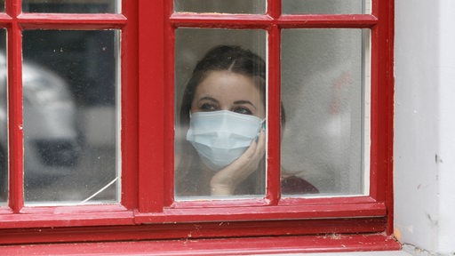 Eine Frau mit Maske steht am Fenster und schaut hinaus (Symbolbild)