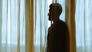 Mann schaut hinter einer Gardine aus dem Fenster (Symbolfoto)
