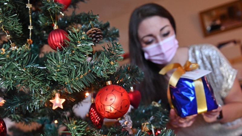 Eine Frau mit Maske sitzt neben einem geschmückten Weihnachtsbaum und nimmt ein Geschenk auf.