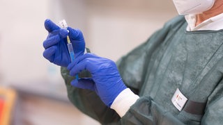 Person mit blauen Gummihandschuhen und Mundschutz zieht eine Spritze mit Impfstoff aus einer Ampulle.