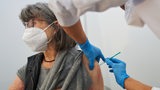 Eine ältere Frau mit grauen schulterlangen Haaren wird von einer Person mit blauen Handschuhen geimpft.