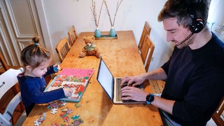 Vater mit Tochter im Homeoffice, er am Computer, das Mädchen mit Puzzle am Esstisch.