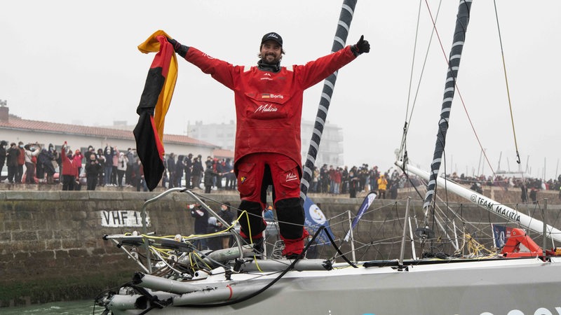 Boris Herrmann steht bei der Zieleinfahrt mit ausgebreiteten Armen und einer Deutschland-Flagge in der Hand an Deck seiner "Seaexplorer".