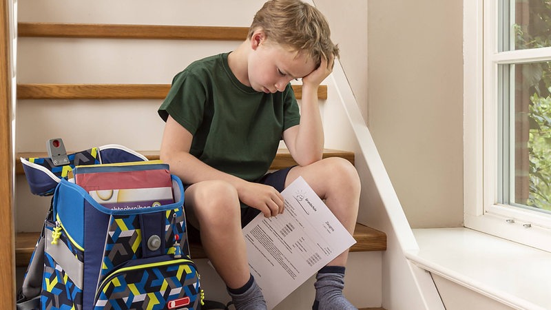 Enttäuschter Junge, 8 Jahre, sitzt zu Hause mit seinem Zeugnis aus der Grundschule auf der Treppe.