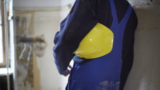 Ein Bauarbeiter steht während der Sanierung eines Altbaus an eine Wand gelehnt und trägt seinen Arbeitsschutzhelm unter dem Arm.
