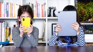 Zwei Schwestern sitzen an einem Tisch, eine hält ein Tablett-PC hoch, die andere ein Smartphone.