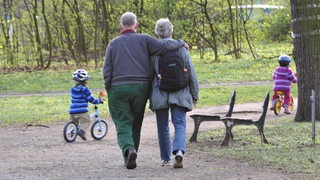Großeltern beim Spaziergang in einem Park. Die Enkelkinder fahren auf Laufrädern voraus. (Symbolbild)