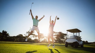 Zwei Golfspieler jubeln auf dem Golfplatz und springen vor Freude in die Luft. Im Hintergund steht ein Golfcart und die Sonne scheint.