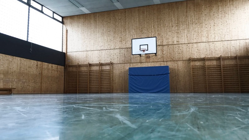 Eine ältere leere Halle mit einem Basketballkorb, unter dem an der Wand eine Turnmatte befestigt ist. 