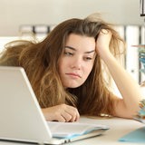 Eine müde blickende Frau mit zerzausten Haaren sitzt vor einem Computer und einem Stapel Akten.