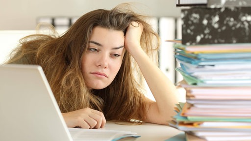 Eine müde blickende Frau mit zerzausten Haaren sitzt vor einem Computer und einem Stapel Akten.