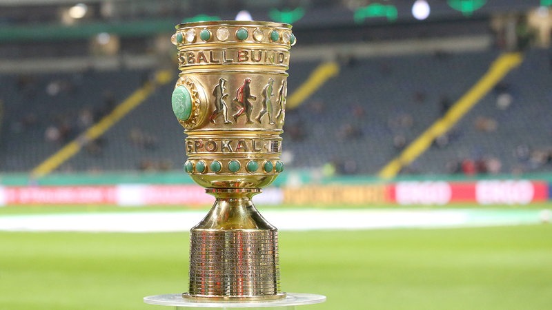 Der goldene DFB-Pokal steht auf einem Plexiglas-Ständer in einem leeren Fußball-Stadion.