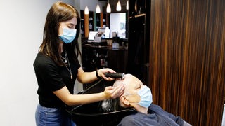 Eine Seniorin erhält eine Haarwäsche von einer Frisuerin, beide tragen dabei eine OP-Maske.