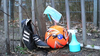 In einem Fußballtor liegen einen Ball, in dem eine Spritze steckt, Fußballschuhe, eine Maske und ein Sprayspender.