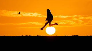 Die Sonne geht am Deich in Bremerhaven unter. Ein Mann ist zu sehen, wie er in die Luft springt.
