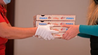 Ein Mann von einem Lieferdienst übergibt mit Handschuhen vier Pizza-Kartons an eine Kundin.