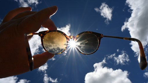 Eine Hand hält eine Sonnenbrille Richtung Himmel der Sonne entgegen.