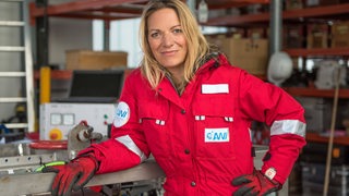 Eine Frau im roten Overall lehnt an einem Gerät.