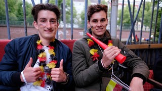 Zwei junge Männer tragen Deutschland-Fankleidung, der linke hebt beide Daumen.