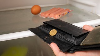 Person hält leere Geldbörse vor leeren Kühlschrank (Symbolbild)