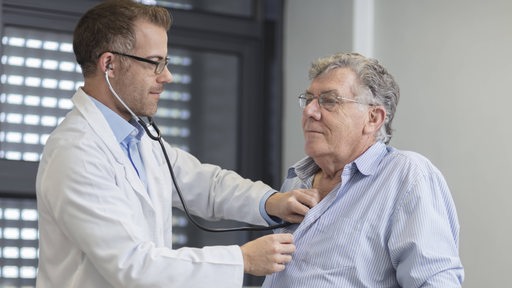 Ein Arzt steht vor einem älteren Patienten und hört den Herzschlag mit einem Stethoskop ab.
