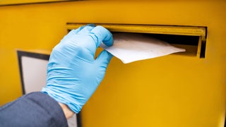 Eine Hand steckt einen Briefumschlag in einen Briefkasten.