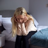 Eine junge Frau sitzt auf dem Bett und wirkt niedergeschlagen (Symbolbild)