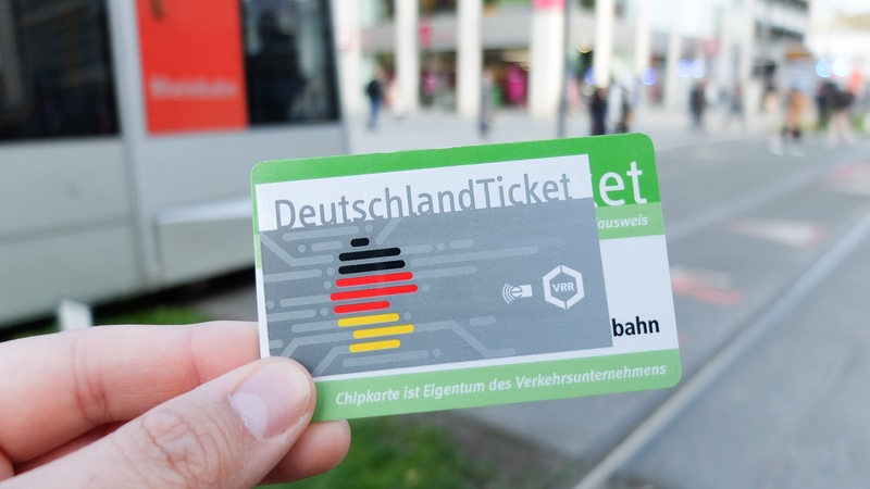 Das bundesweite Deutschlandticket wird als Chipkarte vor einer Tram hochgehalten.