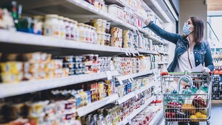 Eine Frau mit Handschuhen und Mundschutz schiebt einen Einkaufswagen durch einen Supermarkt und greift in ein Regal.