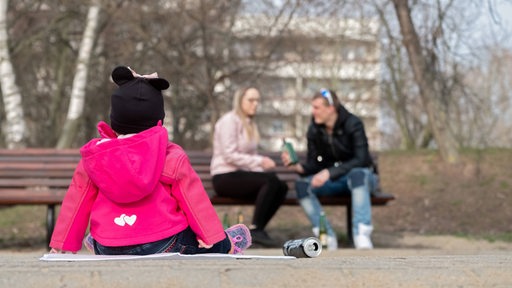 Eltern eines Kindes sitzen auf einer Bank und trinken Bier, während das Kind im Sandkasten spielt (Symbolbild)