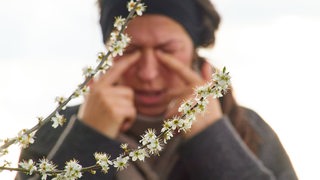 Eine Frau reibt sich die Augen vor einem sprießenden Strauch (Symbolbild)