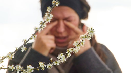 Eine Frau reibt sich die Augen vor einem sprießenden Strauch (Symbolbild)