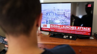 Ein junger Mann schaut Nachrichten über den Krieg in der Ukraine auf einem Fernseher (Symbolbild)