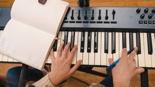 Eine Person spielt auf einem Keyboard und notiert sich Noten (Symbolbild)