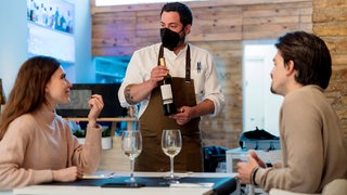 Ein Kellner mit Mund-Nasen-Schutz hält in einem Restaurant eine Flasche Wein.