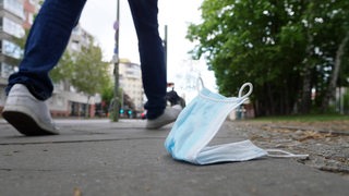 Schutzmaske liegt auf einem Bürgersteig, eine Person geht vorbei (Symbolbild)