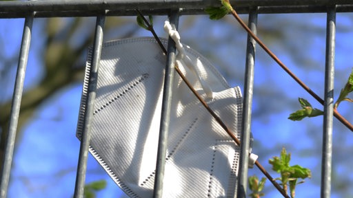Eine benutzte FFP2-Maske hängt an einem Zaun, daneben grüne Triebe eines Busches (Symbolbild)