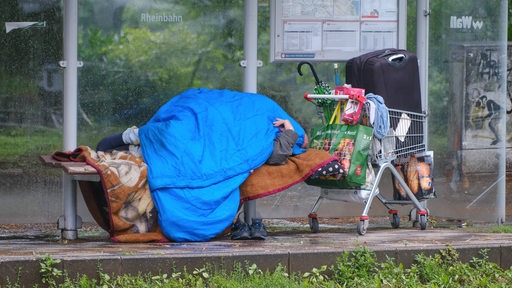 Ein Obdachloser mit Schlafsack bedeckt. Neben ihm ein Einkaufswagen mit seinen Habseligkeiten