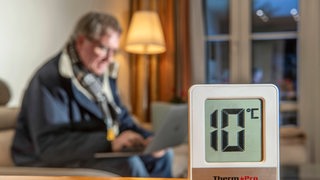 Ein Mann mit Jacke und Schal sitzt auf der Wohnzimmercouch und schaut auf ein Laptop, vorne auf einem Tisch steht ein digitales Thermometer, das 10 Grad anzeigt.