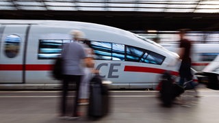 Ein ICE fährt in einen Bahnhof ein, Fahrgäste stehen am Gleis. 