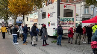 Menschen stehen in einer Schlange an einem großen weißen Bus mit der Aufschrift "Impfen gegen Corona"