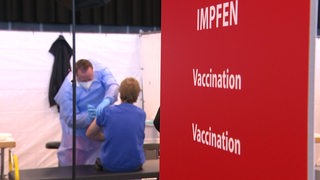 Ein rotes Schild mit der Aufschrift "Impfen, Vaccination, Vaccination". Im Hintergrund wird jemand geimpft.
